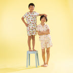 Two children modeling matching short-sleeve pajama sets with a colorful bird print, standing against a solid pale yellow background. The boy stands on a light blue stool, and the girl stands beside him.