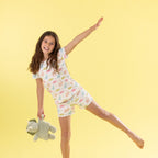 A young girl with long brown hair is captured mid-jump against a solid pale yellow background, wearing a white short-sleeve pajama Set with a colorful pastel dinosaur pattern and holding a green plush toy.
