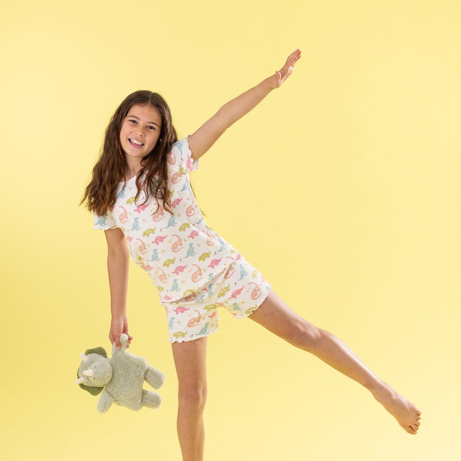A young girl with long brown hair is captured mid-jump against a solid pale yellow background, wearing a white short-sleeve pajama Set with a colorful pastel dinosaur pattern and holding a green plush toy.
