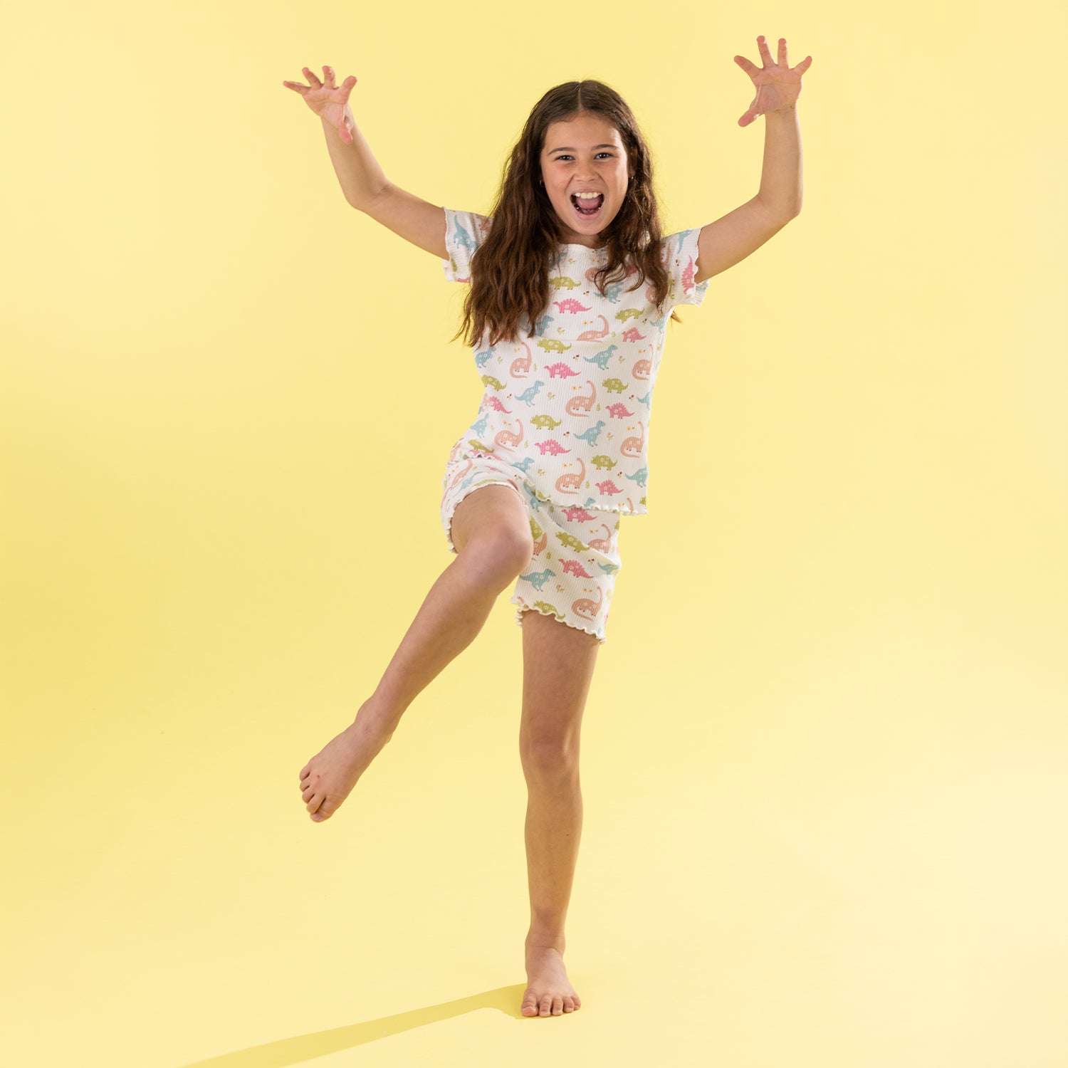 A young girl with long brown hair is captured mid-jump in a playful pose, wearing a matching short-sleeve pajama set with a colorful dinosaur print. She is standing against a solid pale yellow studio background.