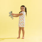A young girl with her hair in a ponytail stands barefoot against a solid pale yellow background, holding a gray plush toy. She is wearing a short-sleeve white nightgown covered in a colorful dinosaur pattern.