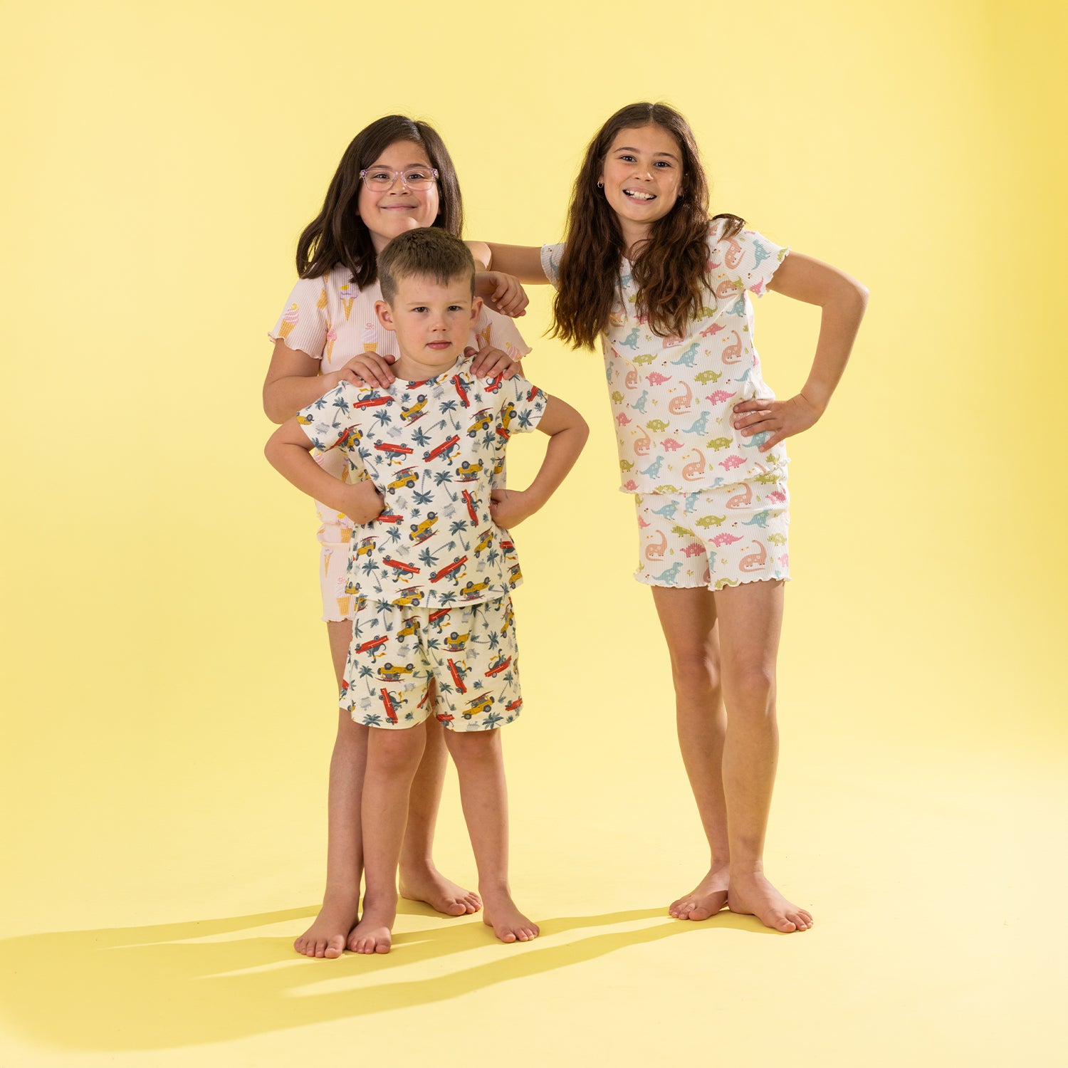 Three children modeling matching short-sleeve pajama sets against a solid pale yellow studio background. The boy in the center wears a white pajama set with a colorful cartoon dinosaur print, while the two girls behind him wear matching white pajama dresses with a pink flamingo print.