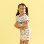 A young girl with dark hair in pigtails stands against a solid pale yellow background, wearing a matching short-sleeve pajama set with a colorful dinosaur print. She is holding a plush green stuffed animal.