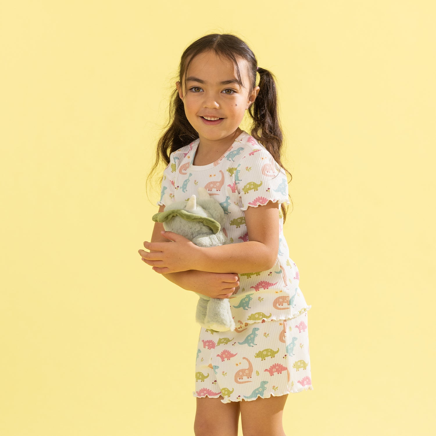 A young girl with dark hair in pigtails stands against a solid pale yellow background, wearing a matching short-sleeve pajama set with a colorful dinosaur print. She is holding a plush green stuffed animal.