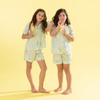 Two girls wearing matching light blue and yellow lemon-patterned pajamas stand barefoot against a solid pale yellow background, each holding a drink with a straw.
