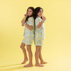 Two girls standing back-to-back wearing matching light green pajama sets with a yellow lemon print, against a solid yellow background.
