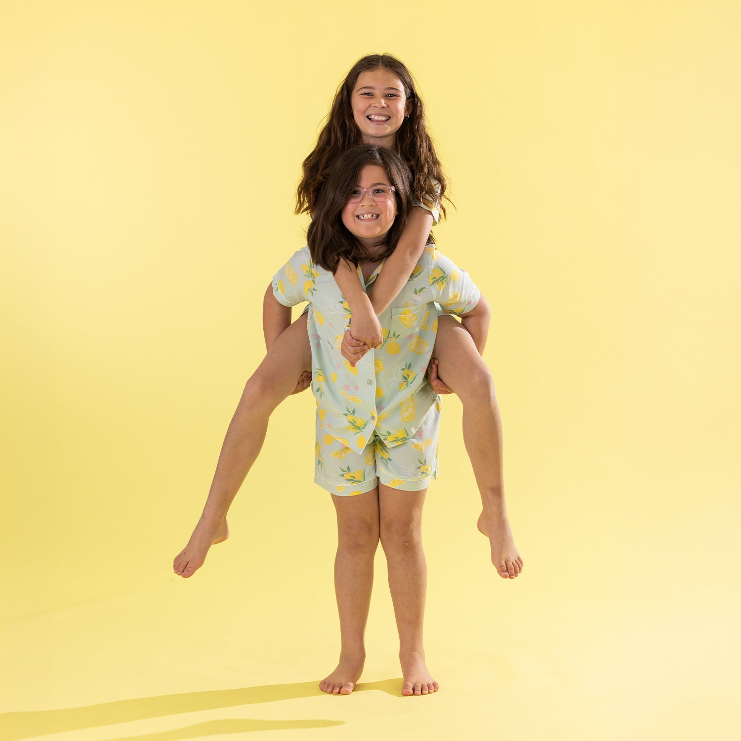 A woman and a girl wearing matching light yellow and green patterned pajamas, with the girl sitting on the woman's shoulders against a solid yellow background.