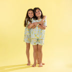 Two young girls wearing matching light green and yellow lemon-print pajamas, standing barefoot against a solid pale yellow background.