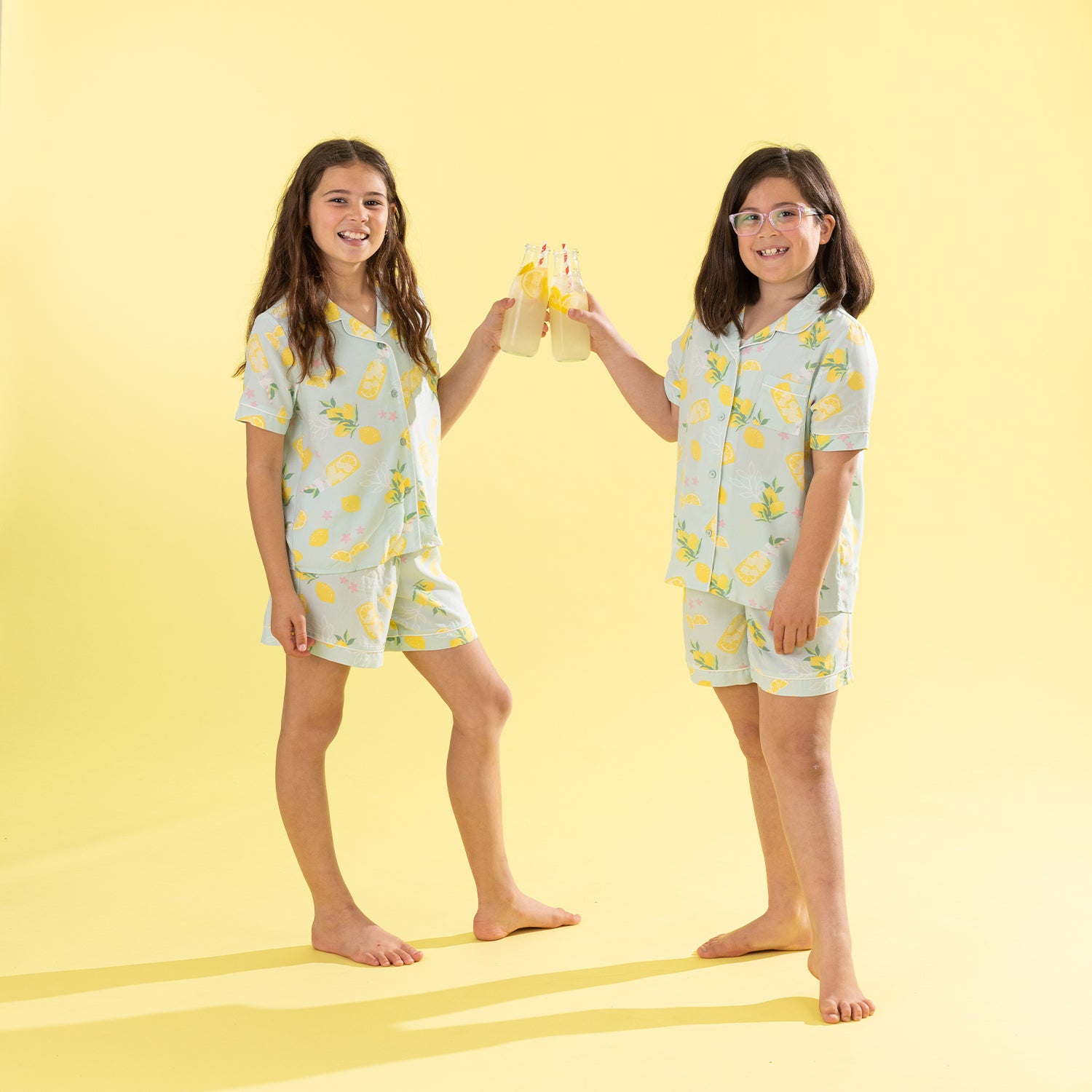 Two young girls standing barefoot against a solid pale yellow background, wearing matching short-sleeve pajama sets with a yellow lemon print. One girl holds a small yellow plush toy.