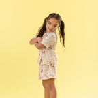A young girl with long brown hair in pigtails poses in a matching short-sleeve pajama set with a colorful seashell & Mermaid pattern, set against a solid pale yellow background.