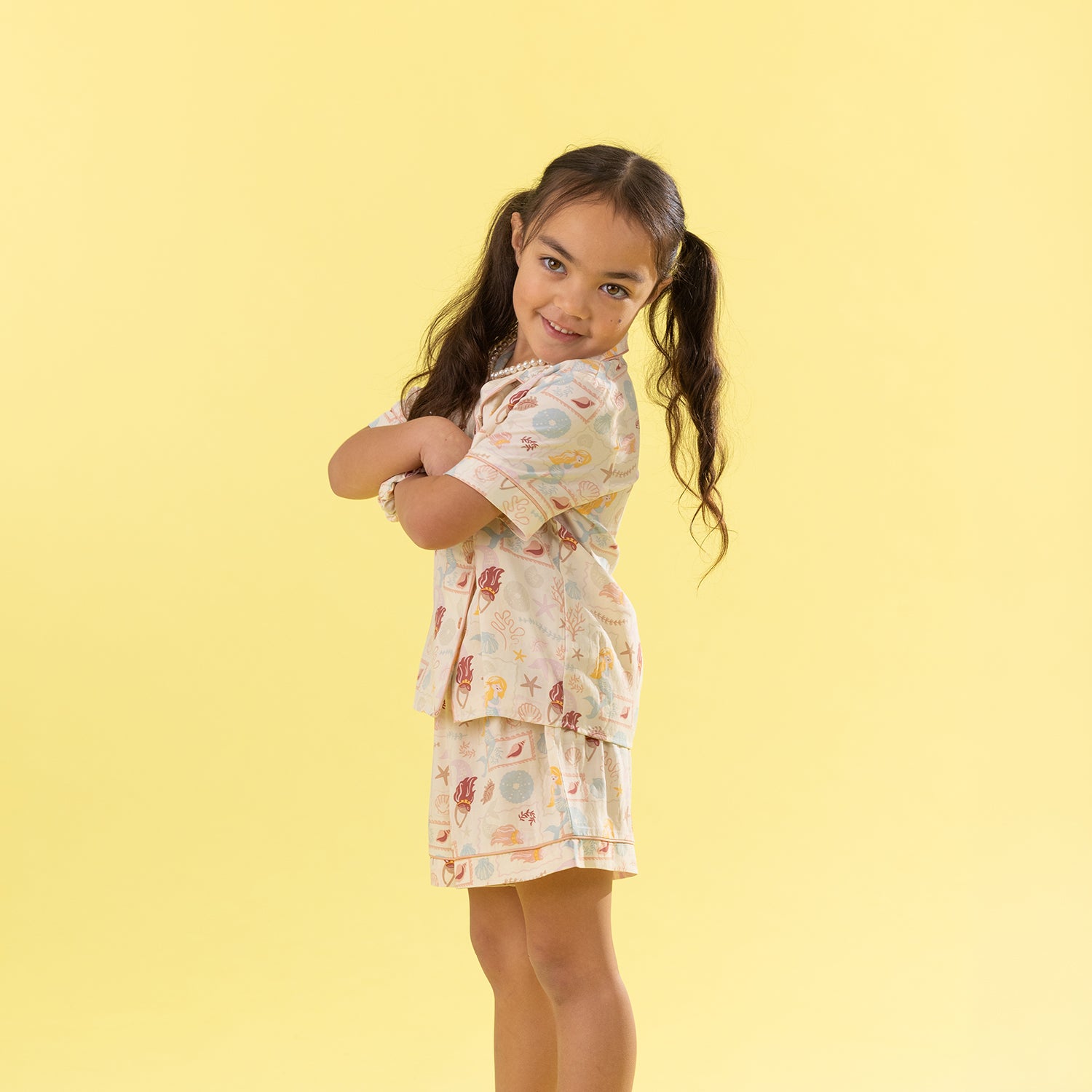 A young girl with long brown hair in pigtails poses in a matching short-sleeve pajama set with a colorful seashell & Mermaid pattern, set against a solid pale yellow background.