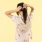 A child viewed from behind wearing a short-sleeve t-shirt with a colorful seashell and starfish print, tying their dark hair in a scrunchie against a solid pale yellow background.