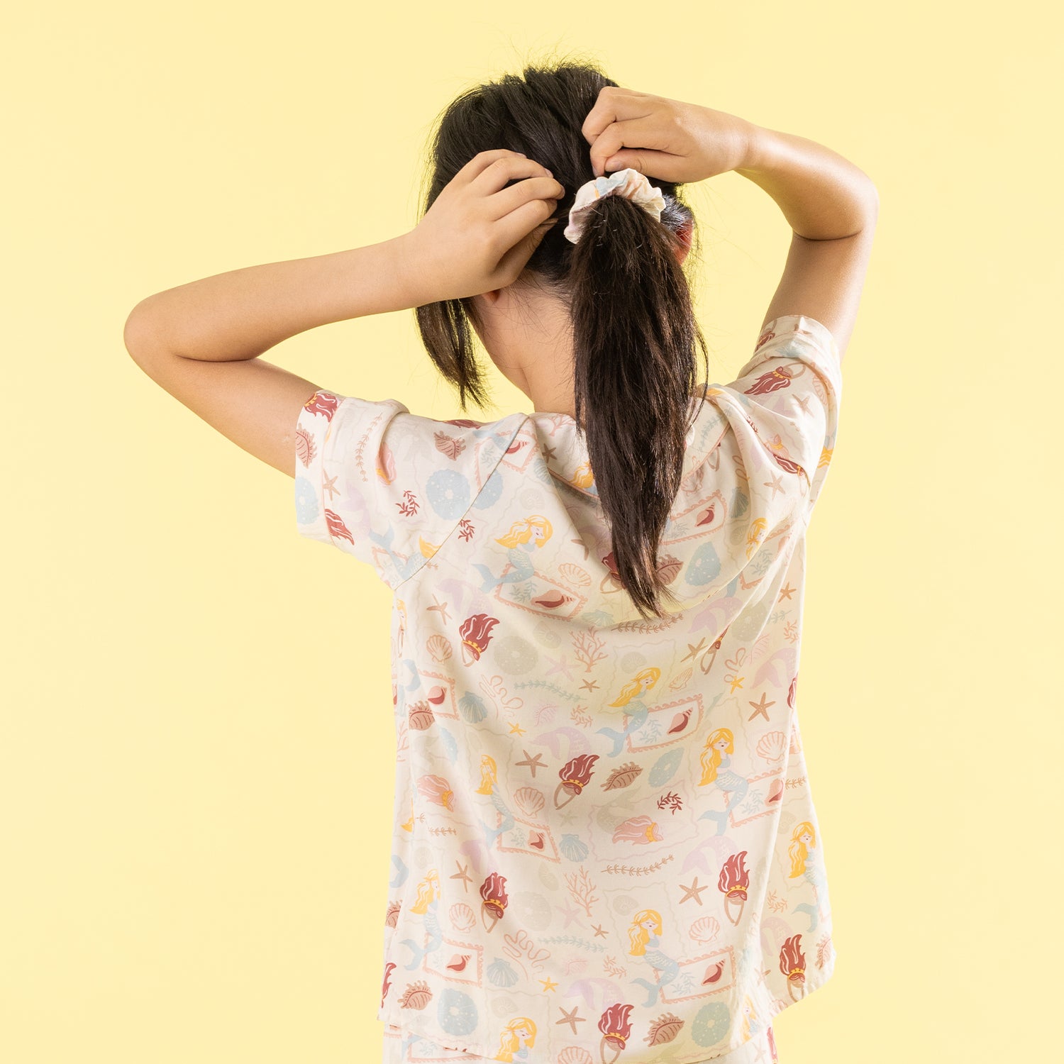 A child viewed from behind wearing a short-sleeve t-shirt with a colorful seashell and starfish print, tying their dark hair in a scrunchie against a solid pale yellow background.