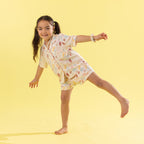 A young girl with dark hair in a ponytail poses dynamically in a matching short-sleeve button-up shirt and shorts pajama set. The pajamas feature a light cream base with a colorful, whimsical print of Mermaids and objects.