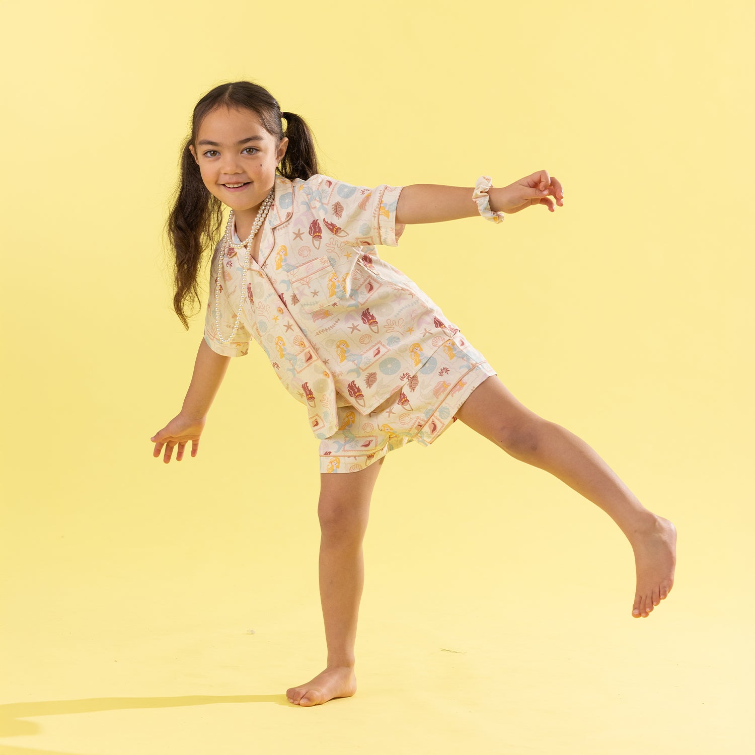 A young girl with dark hair in a ponytail poses dynamically in a matching short-sleeve button-up shirt and shorts pajama set. The pajamas feature a light cream base with a colorful, whimsical print of Mermaids and objects.