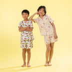 Two children modeling matching short-sleeve pajama sets against a solid pale yellow background. The boy wears a white pajama set with a colorful Dino print, and the girl wears a light beige pajama set with a colorful Mermaid print.