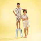 Two children modeling matching short-sleeve pajama sets with a colorful Dino print on a white background, standing against a solid yellow backdrop. The child on the left stands on a light blue stool, and the child on the right stands beside it.