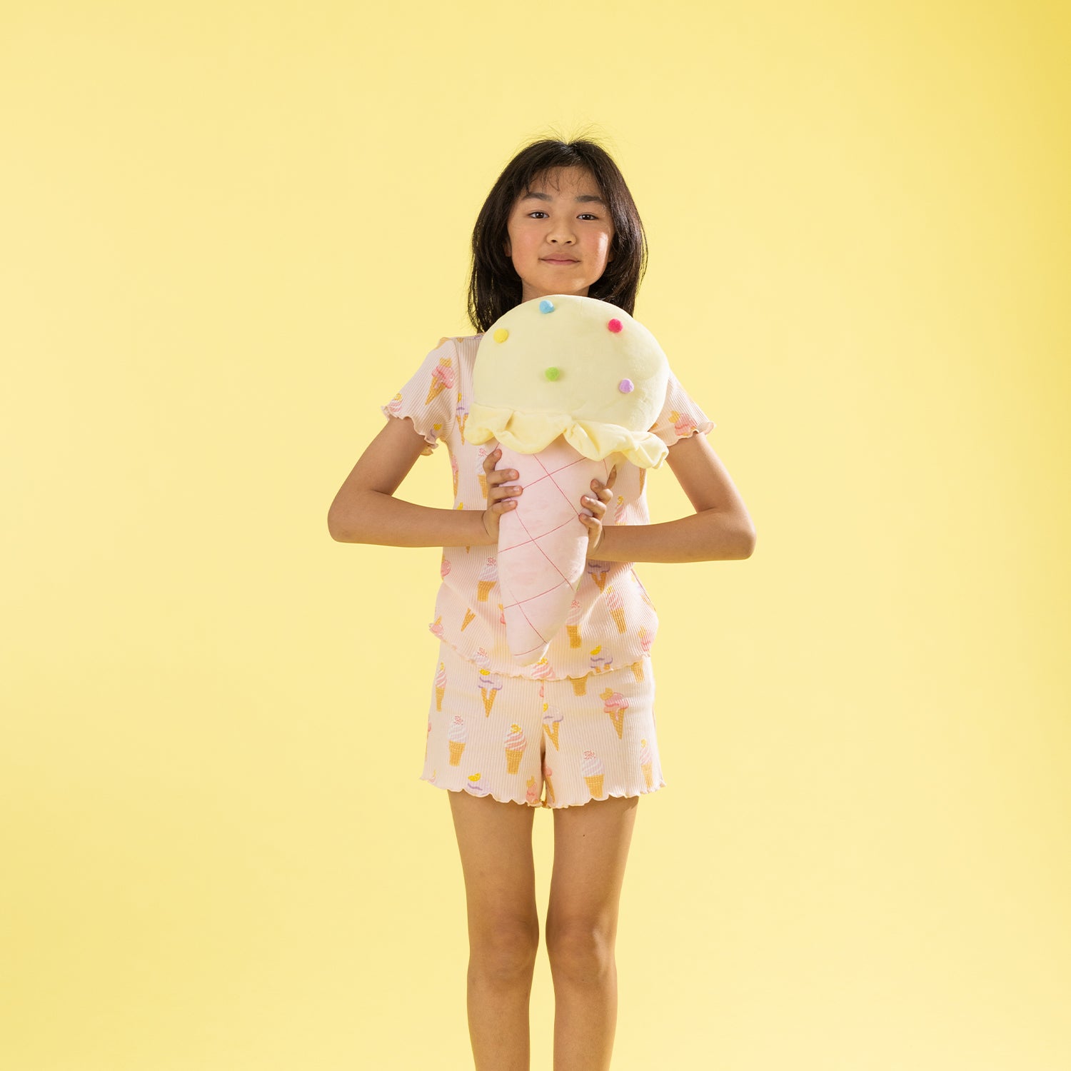 A young girl stands against a solid pale yellow background, holding a large plush ice cream cone toy. She is wearing a light pink pajama set with a small ice cream cone pattern.