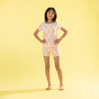 A young girl stands with hands on hips wearing a two-piece pajama set featuring a white base with a yellow ice cream cone pattern, set against a solid yellow studio background.