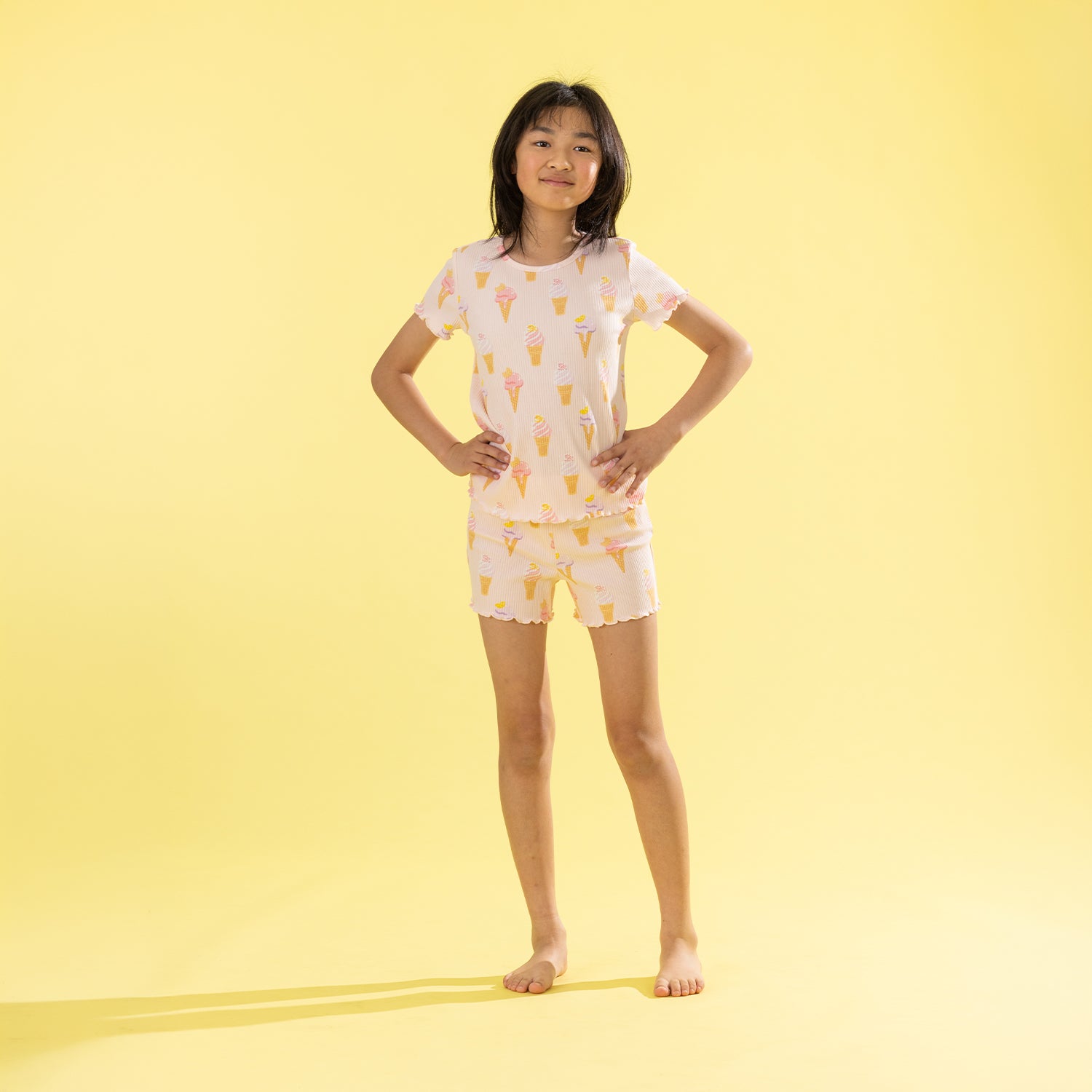 A young girl stands with hands on hips wearing a two-piece pajama set featuring a white base with a yellow ice cream cone pattern, set against a solid yellow studio background.