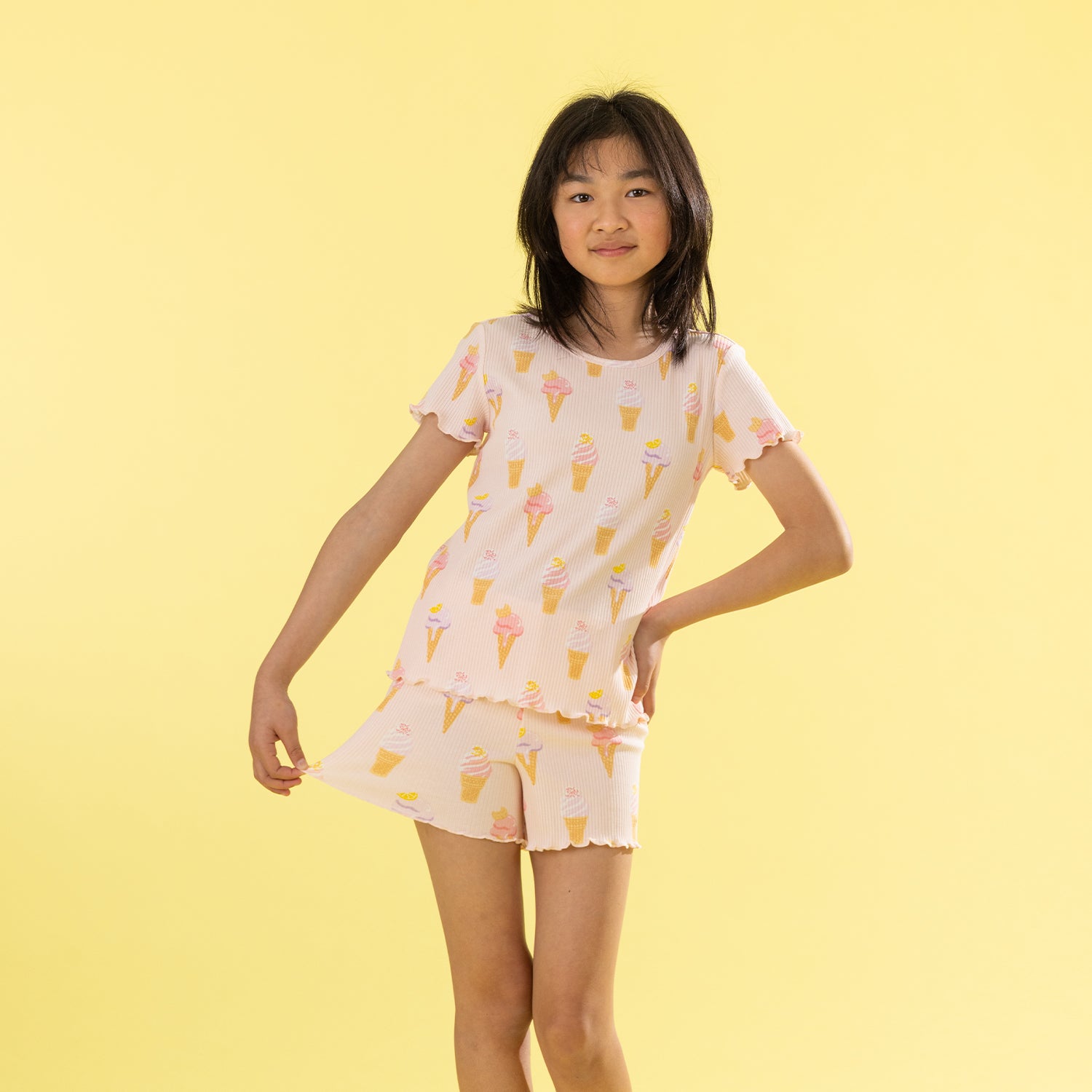 A young girl modeling a two-piece pajama set featuring a light pink base with a yellow and orange ice cream cone pattern, set against a solid pale yellow background.