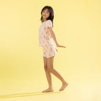 A young girl stands barefoot against a solid pale yellow background, wearing a two-piece pajama set with a white base and a colorful ice cream cone pattern.