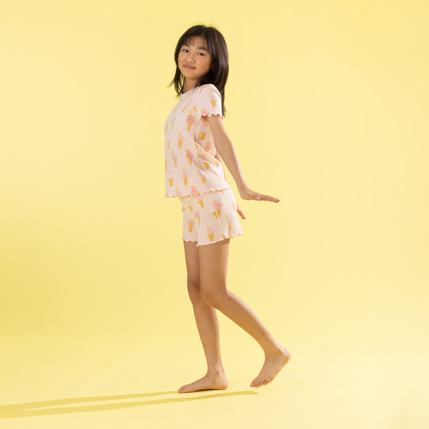A young girl stands barefoot against a solid pale yellow background, wearing a two-piece pajama set with a white base and a colorful ice cream cone pattern.
