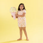 A young girl with glasses stands barefoot against a solid pale yellow background, holding a large plush ice cream cone toy. She is wearing a light pink short-sleeve pajama set with a yellow and orange ice cream cone pattern.