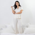 A woman modeling a two-piece pajama set consisting of a short-sleeve button-up shirt and matching wide-leg pants, both in a warm off-white or cream color. She is standing on a bed with white pillows and bedding against a clean white wall.