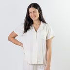 A woman modeling a short-sleeve button-front pajama shirt in a warm off-white or cream color, paired with matching off-white pants, against a plain light gray studio background.