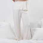 A person wearing a matching cream-colored pajama set with a short-sleeve button-front top and wide-leg pants, standing on a white bed with textured pillows.