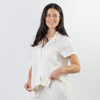 A woman with long dark hair wearing a white short-sleeve button-up shirt and matching white trousers, posing against a solid white studio background.