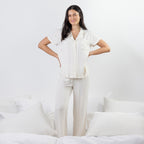 A woman modeling a matching short-sleeve button-front pajama top and wide-leg pajama pants set in a bright white bedroom setting.