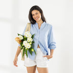 Model wearing the Cove Breeze Stitch Shorts in Ivory with a light blue shirt, styled with a woven shoulder bag filled with white flowers and a baguette.