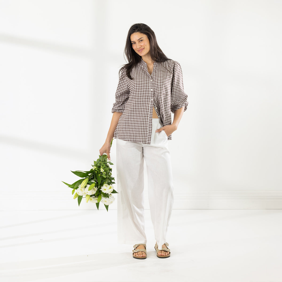 Full-length view of the model wearing the Cove Breeze Collarless Shirt in Chocolate Check with ivory pants, holding a bouquet of white flowers.