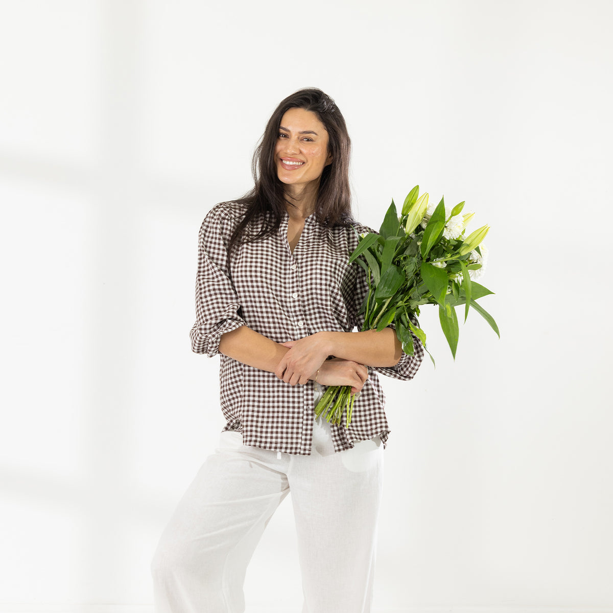 Model wearing the Cove Breeze Collarless Shirt in Chocolate Check with ivory pants, holding a bouquet of white flowers.