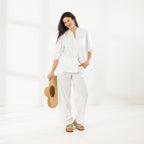 Full-length view of the model wearing the Cove Breeze Collarless Shirt in Ivory with coordinating ivory pants, holding a wide-brim straw hat.
