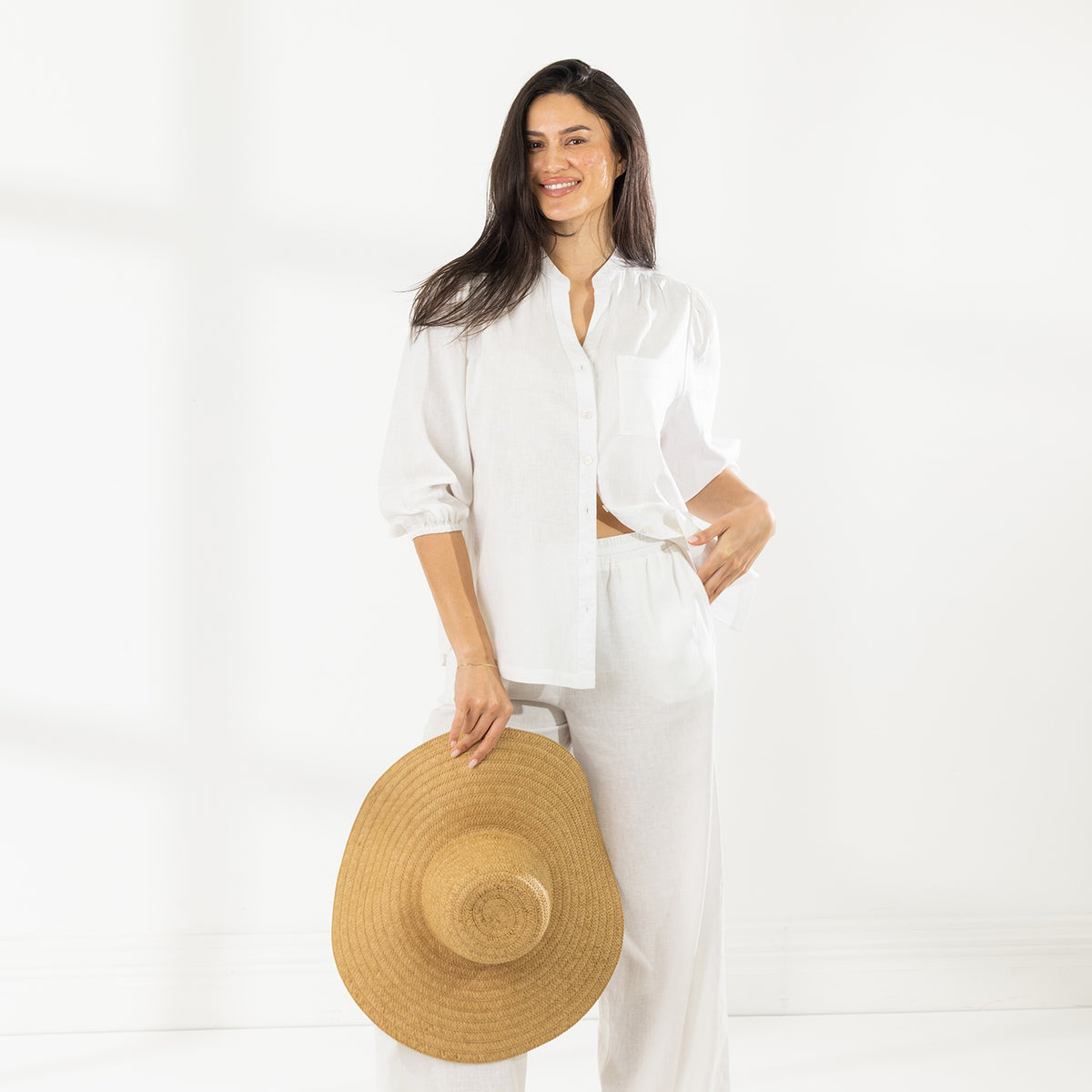 Full-length view of the model wearing the Cove Breeze Collarless Shirt in Ivory with coordinating ivory pants, holding a large straw sunhat.