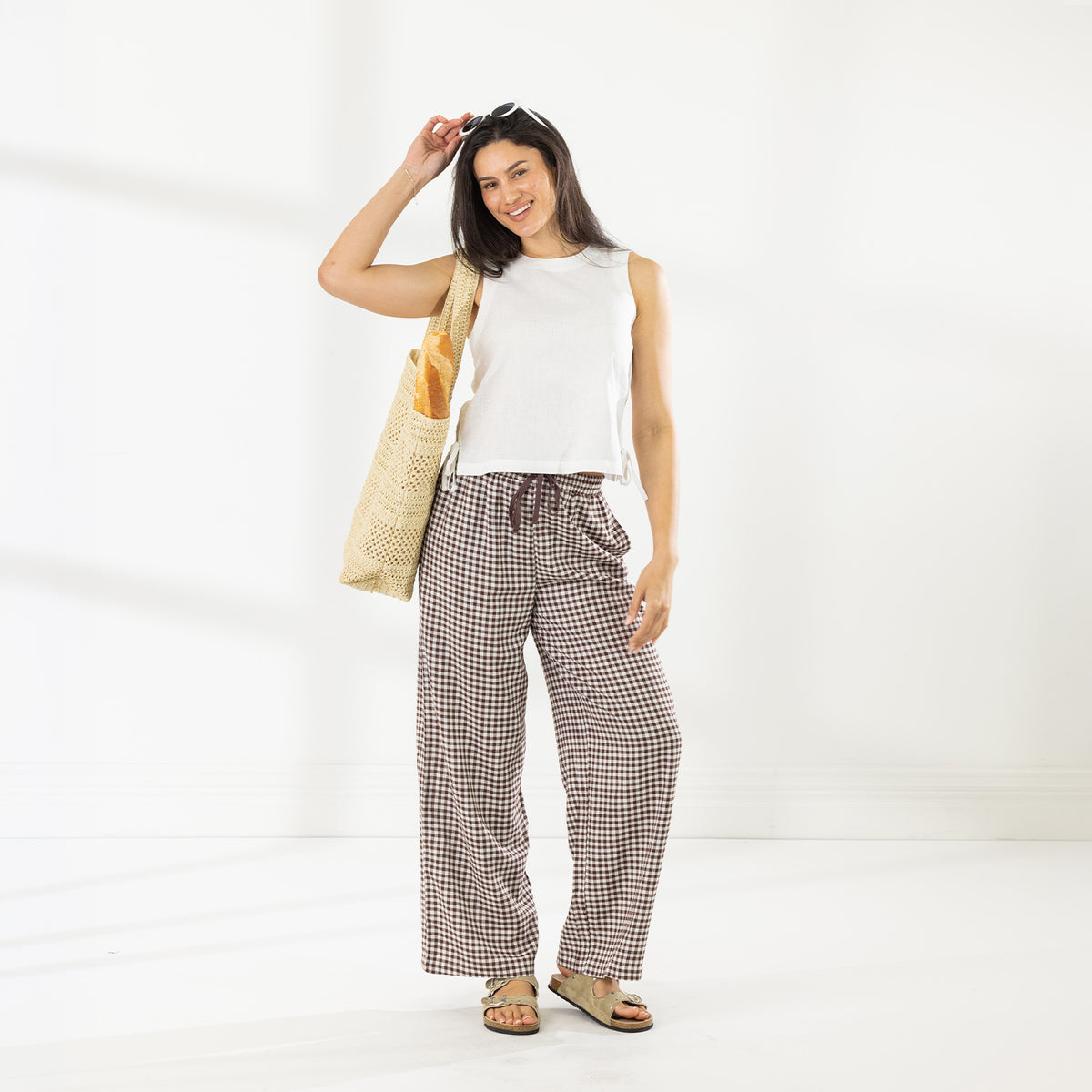 Full-length view of the Cove Wide Leg Pants in Chocolate styled with an ivory sleeveless top and woven tote bag.
