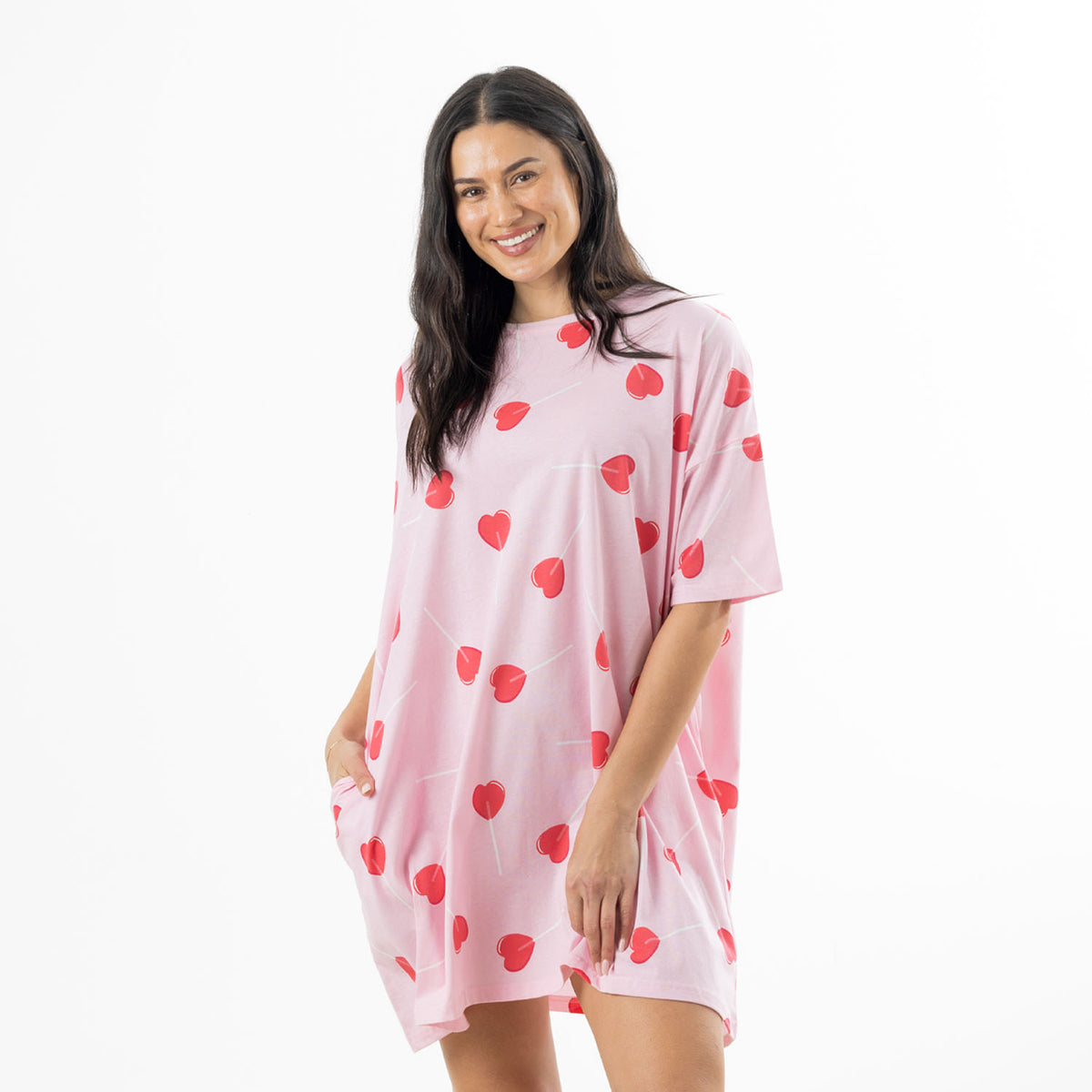 Woman modelling a soft pink oversized sleep tee with an all-over red heart-shaped lollipop print and short sleeves against a white background.