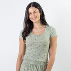 A woman with long dark hair wearing a sage green pj top with a subtle tonal floral pattern, photographed against a plain light gray studio background.