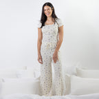 A woman stands on a bed wearing a matching short-sleeve top and pants pajama set in cream with a small blue floral print. The room has white bedding and pillows against a white wall.