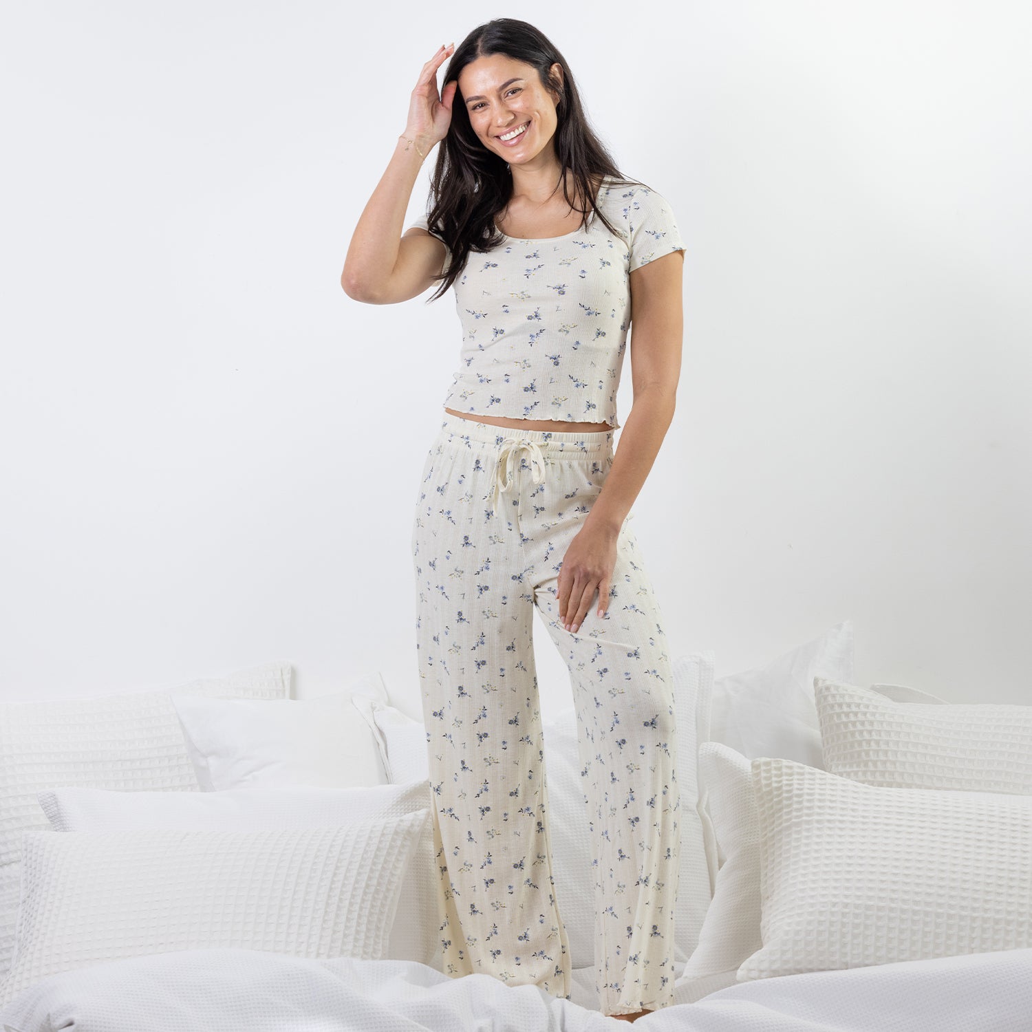 A woman stands on a bed wearing a matching two-piece pajama set with a small blue floral print on a warm off-white background. The set includes a short-sleeve crop top and wide-leg pants with a drawstring waist.