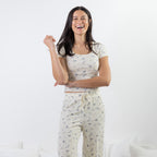 A woman modeling a matching two-piece pajama set consisting of a short-sleeve crop top and drawstring pants, both in a cream color with a small blue floral print, against a white studio background.