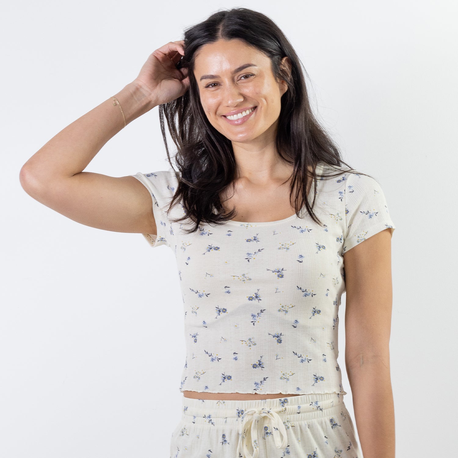 A woman modeling a short-sleeve scoop-neck crop pj top with a small blue and yellow floral print on a cream background, paired with matching cream drawstring shorts, against a plain white studio background.