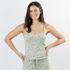 A woman modeling a light sage green pajama set with a subtle floral pattern, featuring a camisole top and matching shorts, against a plain light gray background.