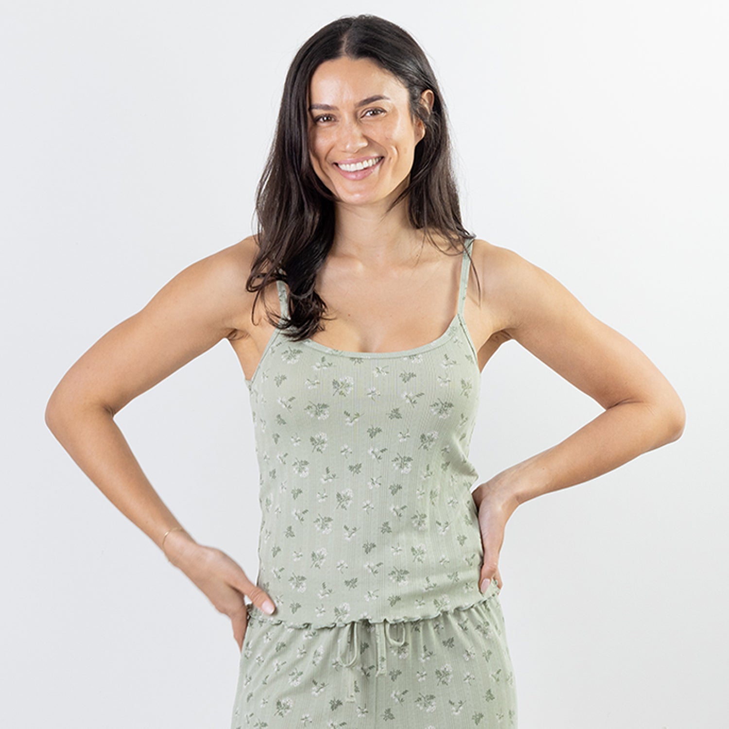 A woman modeling a light sage green pajama set with a subtle floral pattern, featuring a camisole top and matching shorts, against a plain light gray background.