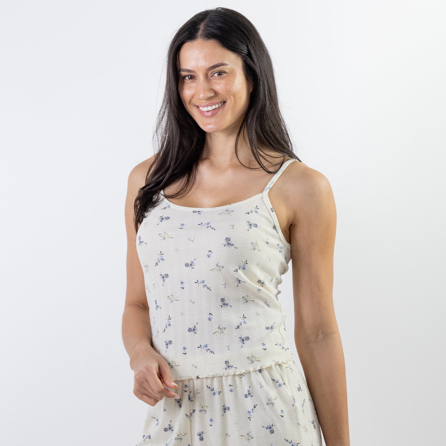 A woman with long dark hair wearing a cream-colored spaghetti-strap pj cami with a small blue and yellow floral print, photographed against a solid white studio background.