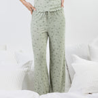 A person wearing light sage green drawstring pj pants with a small floral print, standing in a bright bedroom with white bedding and pillows.