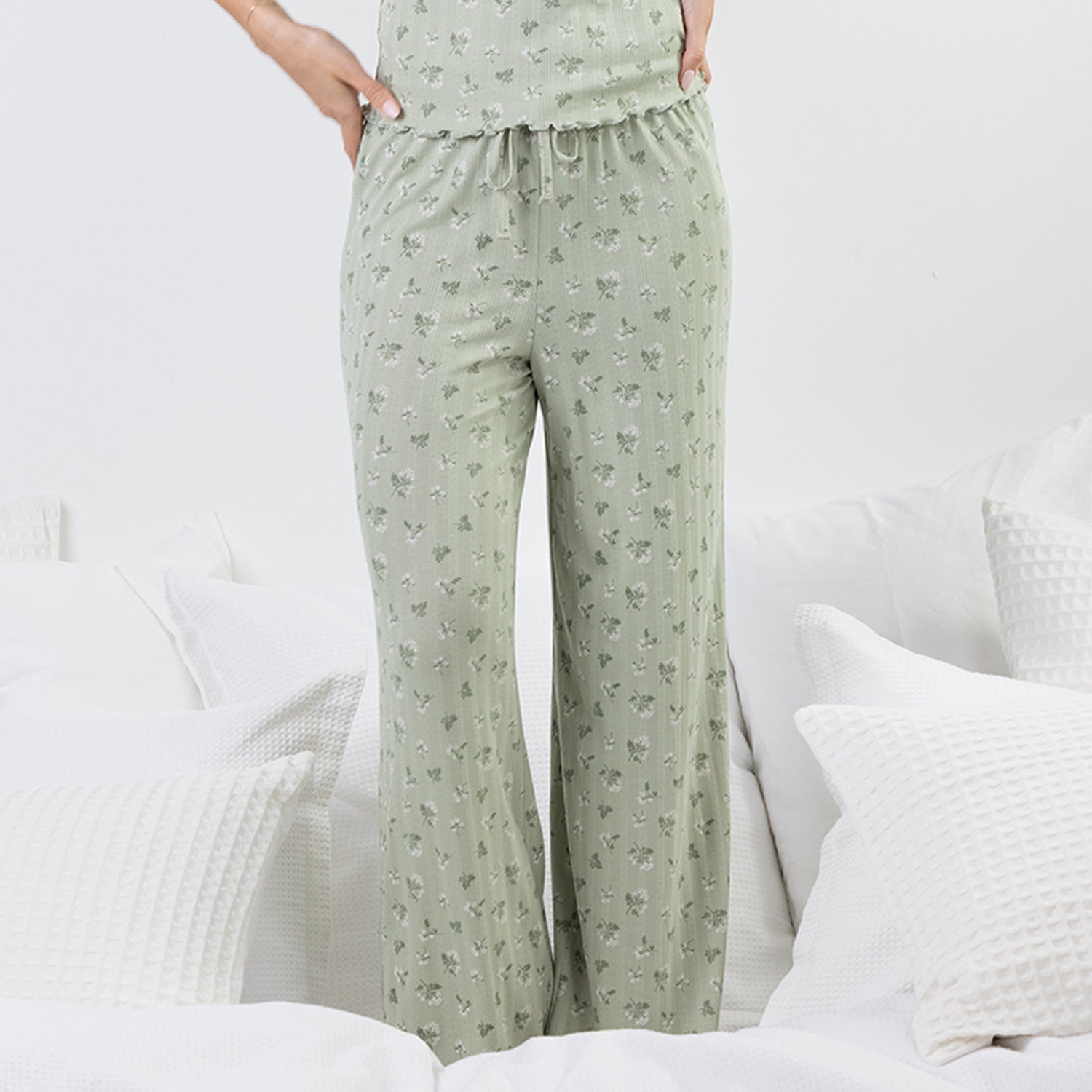 A person wearing light sage green drawstring pj pants with a small floral print, standing in a bright bedroom with white bedding and pillows.
